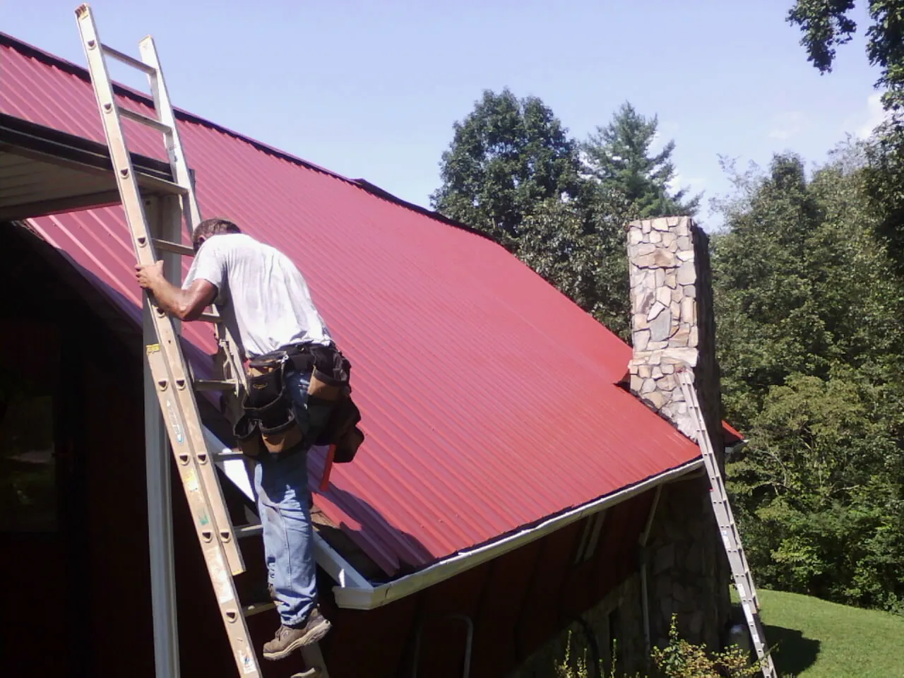 Roofer on a ladder beside a red metal roof and stone chimney — Soffit & Fascia Repair by Roofing Brothers in Robeline